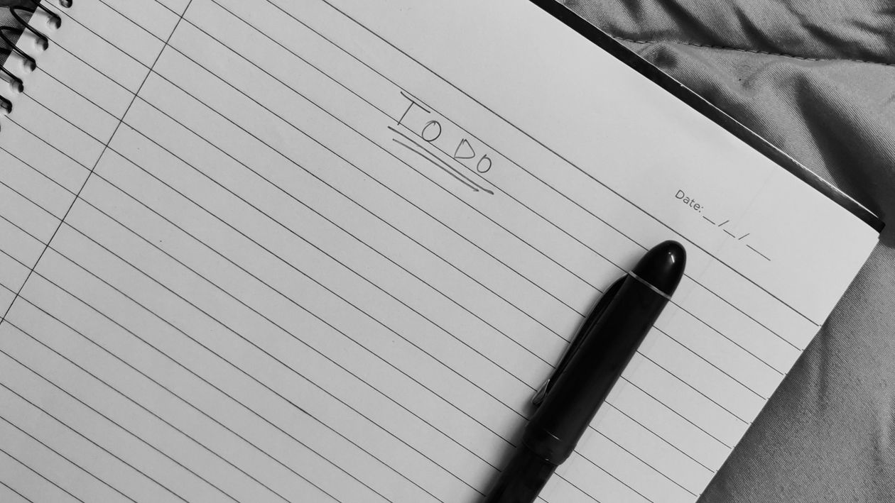A real photograph of a notebook listing debts in order, with a pen resting on the page